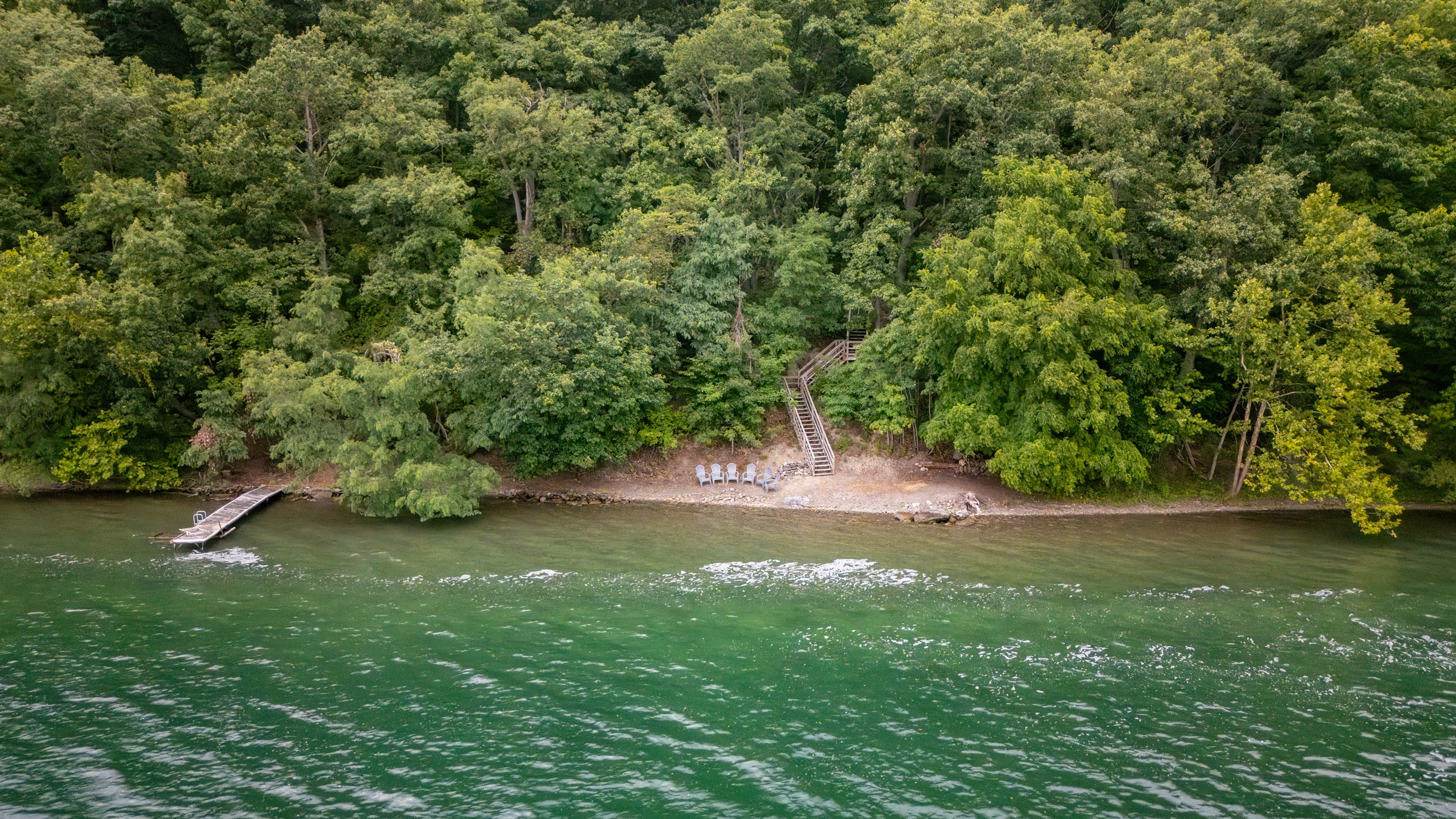 Aerial view of a private, wooded shoreline retreat with direct lake access, dock, and Adirondack chairs for lakeside relaxation.