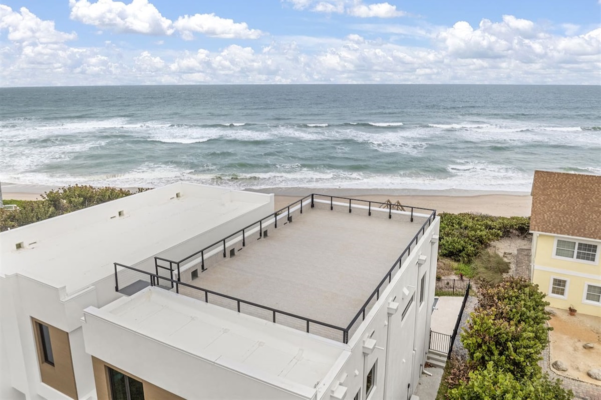 Captured by drone, the rooftop offers a stunning view of the neighborhood's houses, with the ocean stretching beyond and lush greenery surrounding the area, creating a peaceful and scenic escape.