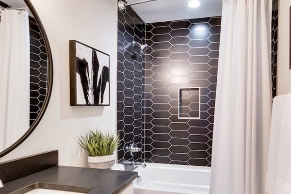 - Striking black tile shower with geometric pattern and built in niche for essentials - Round mirror and dark vanity top add contrast and modern flair to the space - Bright lighting, framed art, and greenery finish the spa like look with style