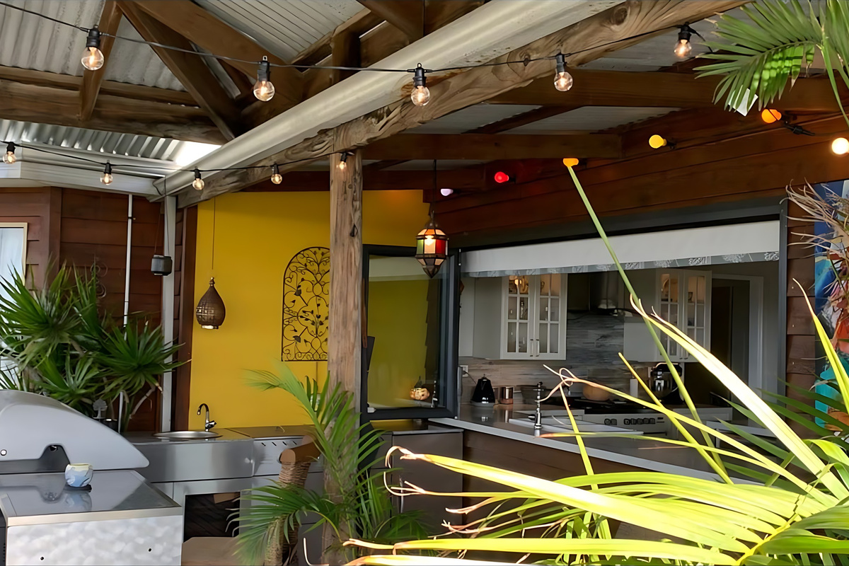 The outdoor kitchen beside the gazebo deck has a stainless steel BBQ, bench space and a sink under festoon lights, with a bifold window opening straight through to the indoor kitchen.