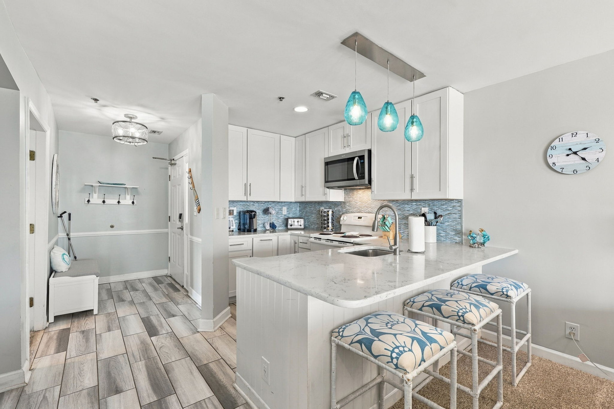 Kitchen counter bar seating with three patterned barstools, quartz countertops, white shaker cabinets, mosaic backsplash, and blue pendant lighting adjacent to the entryway.