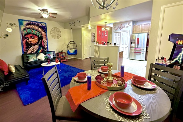 This shared area is vibrant and eclectic with bold decor, including a colorful mural & hanging chair. The dining area has a wooden table set with red dishware, candles, & a gold centerpiece.