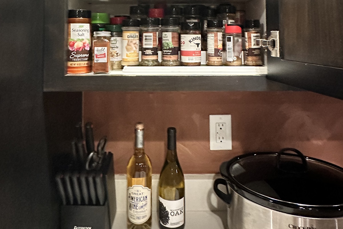 The image shows a cluttered kitchen shelf containing various spice jars, including seasoning salt and herbs, above a countertop with two bottles of white wine, a set of knives in a black holder, and a black crock pot.