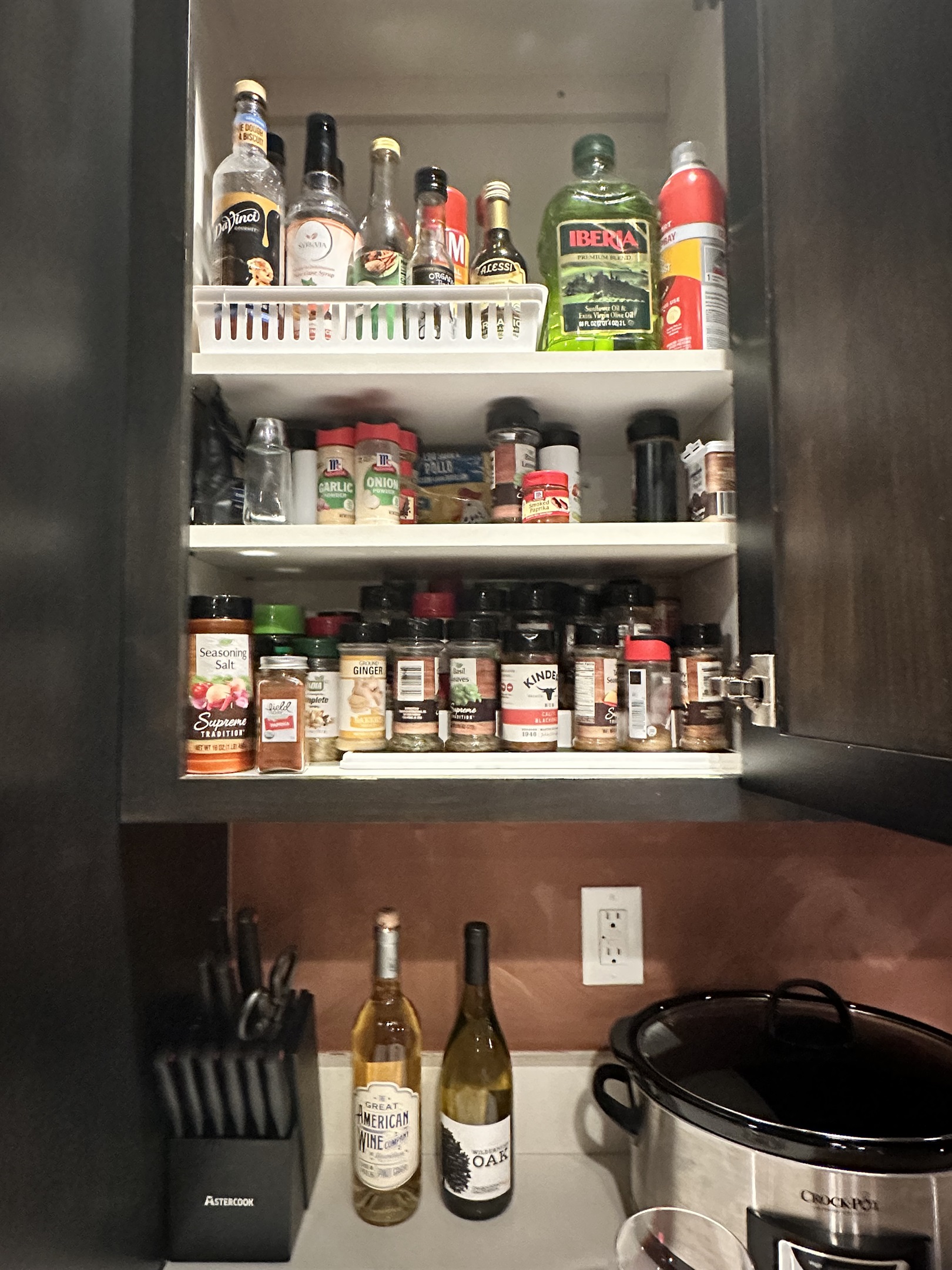 A kitchen cabinet shelf filled with various bottles of sauces and oils, alongside numerous spice jars and seasonings, while a wine rack with two bottles of white wine and a black slow cooker sit on the countertop below.