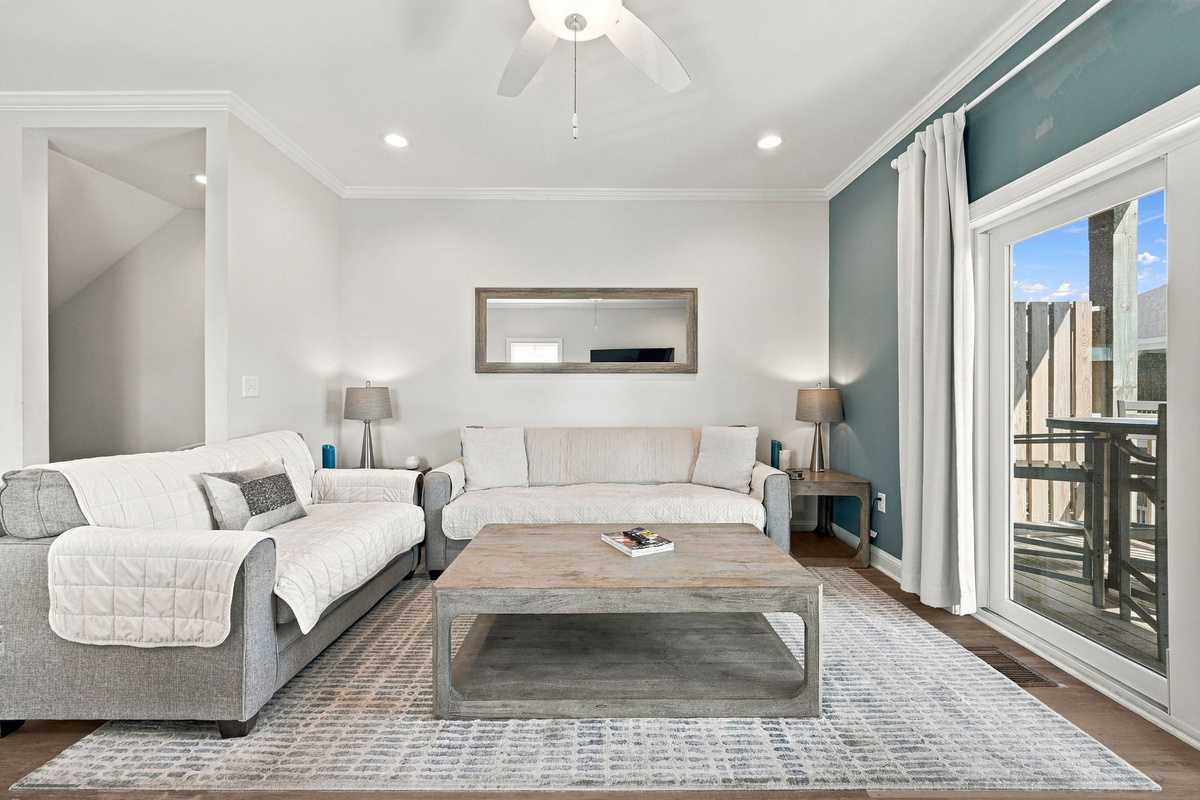 Bright, coastal-inspired living room featuring comfortable plush seating, a rustic wood coffee table, recessed lighting, and a sliding glass door opening directly onto a private deck setup.   