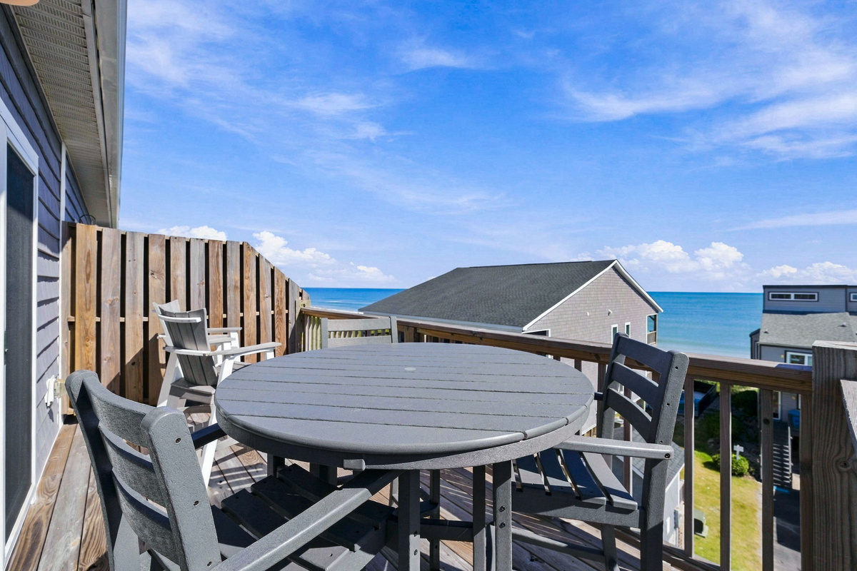 Spacious upper-level private deck featuring a round dining table with seating, Adirondack chairs, a privacy wall, and expansive open-air views looking out toward the ocean.