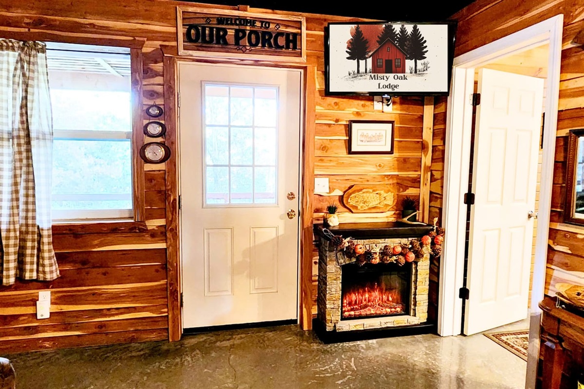 The heart of the lodge welcomes you with a glowing electric fireplace, rustic wood walls, and vintage cabin decor. Natural light spills through the gingham-curtained window, setting the tone for warm, relaxing days at Misty Oak Lodge.