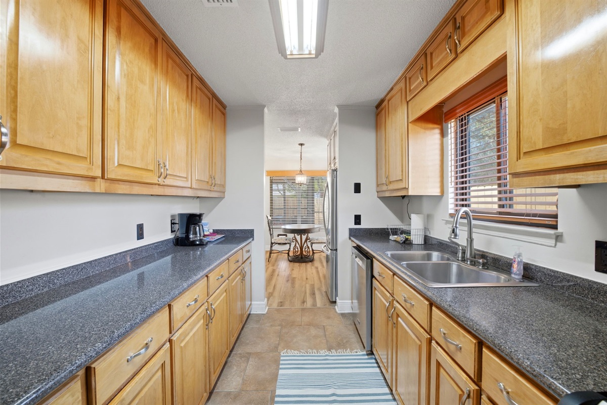 This bright galley-style kitchen offers generous counter space on both sides, making meal prep easy and convenient. Large cabinets, stainless steel appliances, and a window above the sink create a practical and inviting workspace.