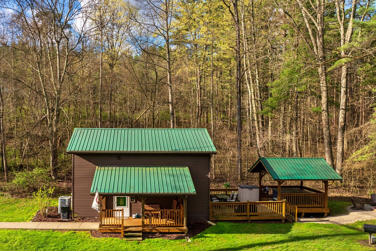 This exterior picture shows the living space, featuring a hot tub on the deck to the right, with a firepit and charcoal grill situated in the yard.