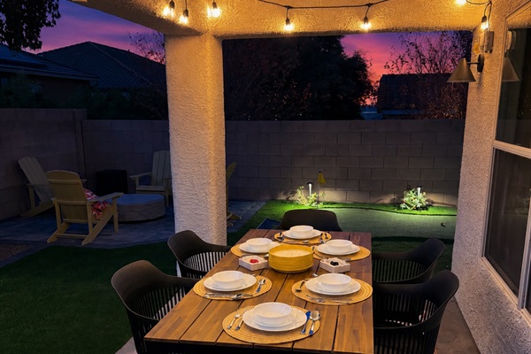Covered outdoor 6-person dining area with soft string lighting and our firepit beyond, at night. -Adventure Awaits.