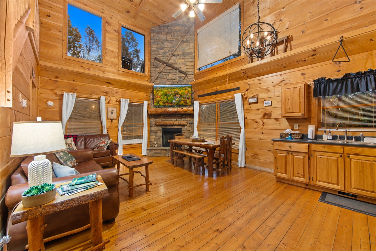 This open-concept cabin living area boasts cozy leather couches, a rustic wooden staircase, and a fully equipped kitchen with black appliances, all set within a warm, wood-paneled interior.