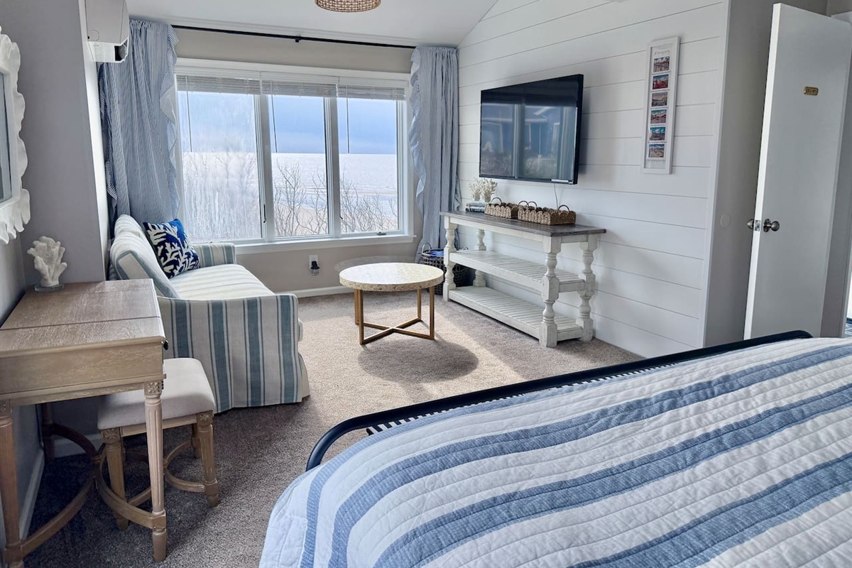 Enjoy a serene coastal retreat featuring a comfortable sitting area with a striped sofa, a scenic window view, and a wall-mounted television, all designed to provide a perfect, relaxing environment for your vacation.