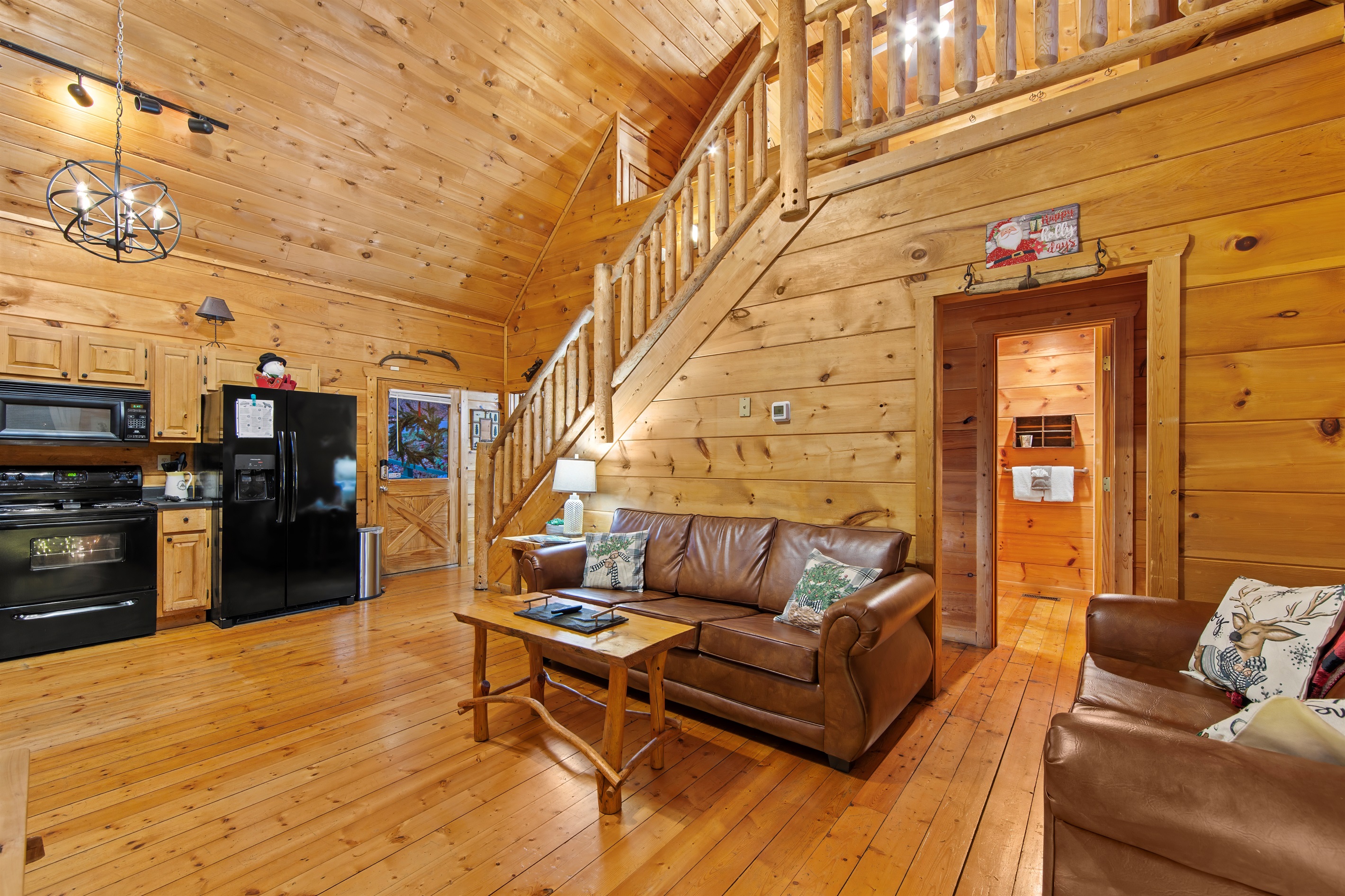 This open-concept cabin living area boasts cozy leather couches, a rustic wooden staircase, and a fully equipped kitchen with black appliances, all set within a warm, wood-paneled interior.