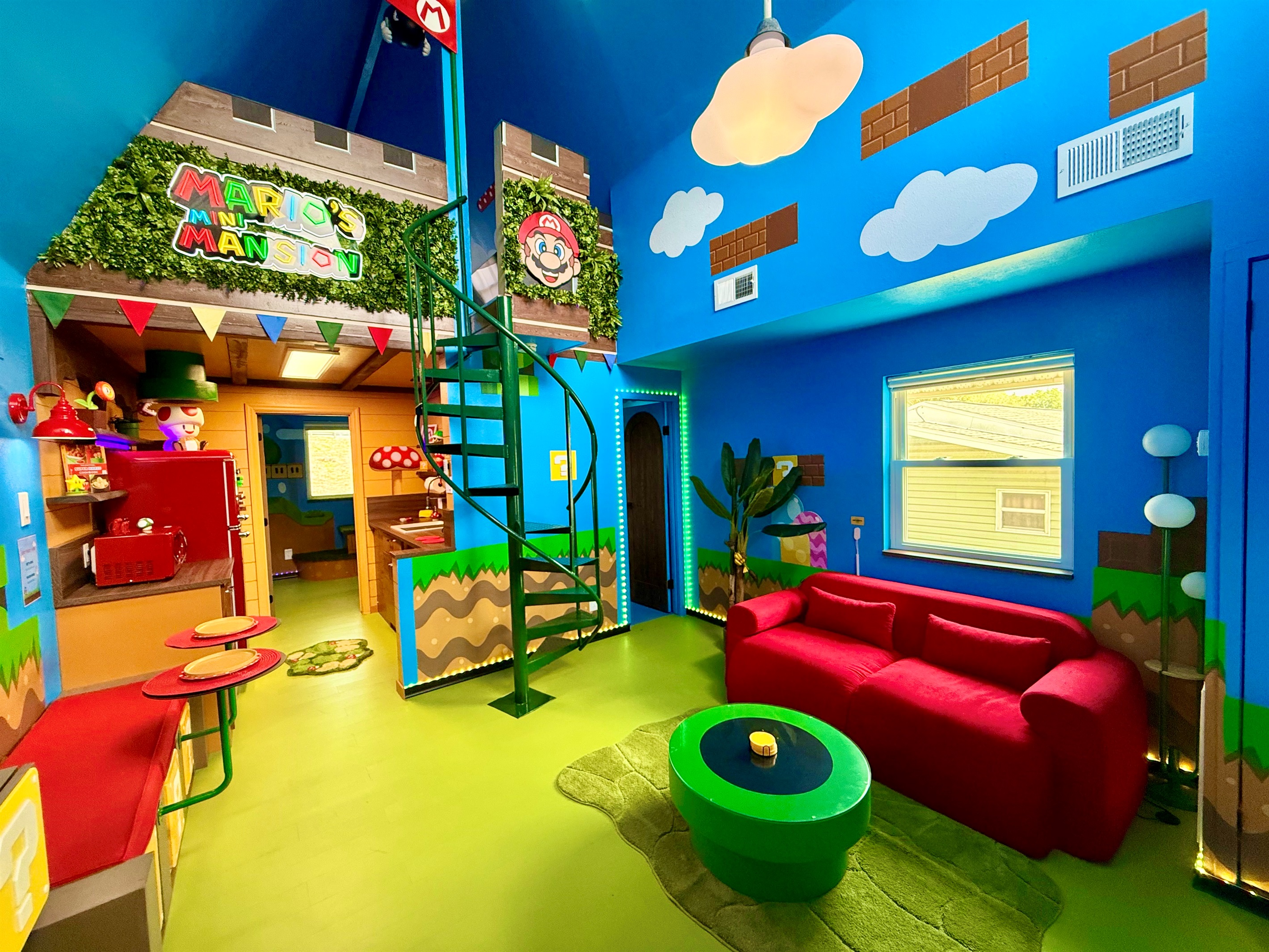 Explore the "castle" area with spiral staircase to the lofted Mario's Mini Mansion king bed! This vibrant blue living room features a squishy red sofa, a green coffee table, custom signage, and playful Mario details -- endless adventures for all.
