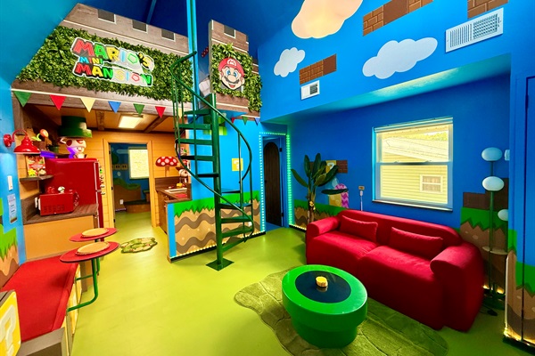 Explore the "castle" area with spiral staircase to the lofted Mario's Mini Mansion king bed! This vibrant blue living room features a squishy red sofa, a green coffee table, custom signage, and playful Mario details -- endless adventures for all.