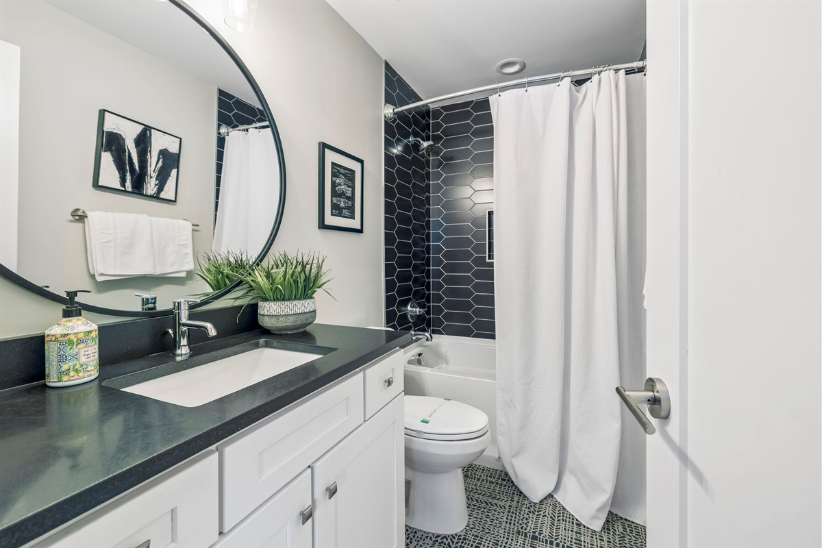 Striking black tile shower with geometric pattern and built in niche for essentials - Round mirror and dark vanity top add contrast and modern flair to the space - Bright lighting, framed art, and greenery finish the spa like look with style