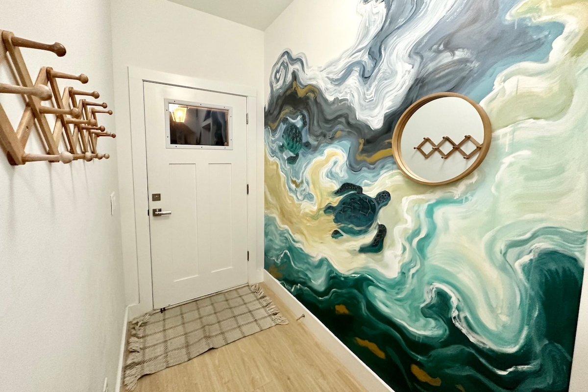 You come in through this. Hooks for wet towels on one side, a hand-painted sea turtle riding the surf on the other, and the whole house opening up beyond it. Guests photograph this wall before they unpack.