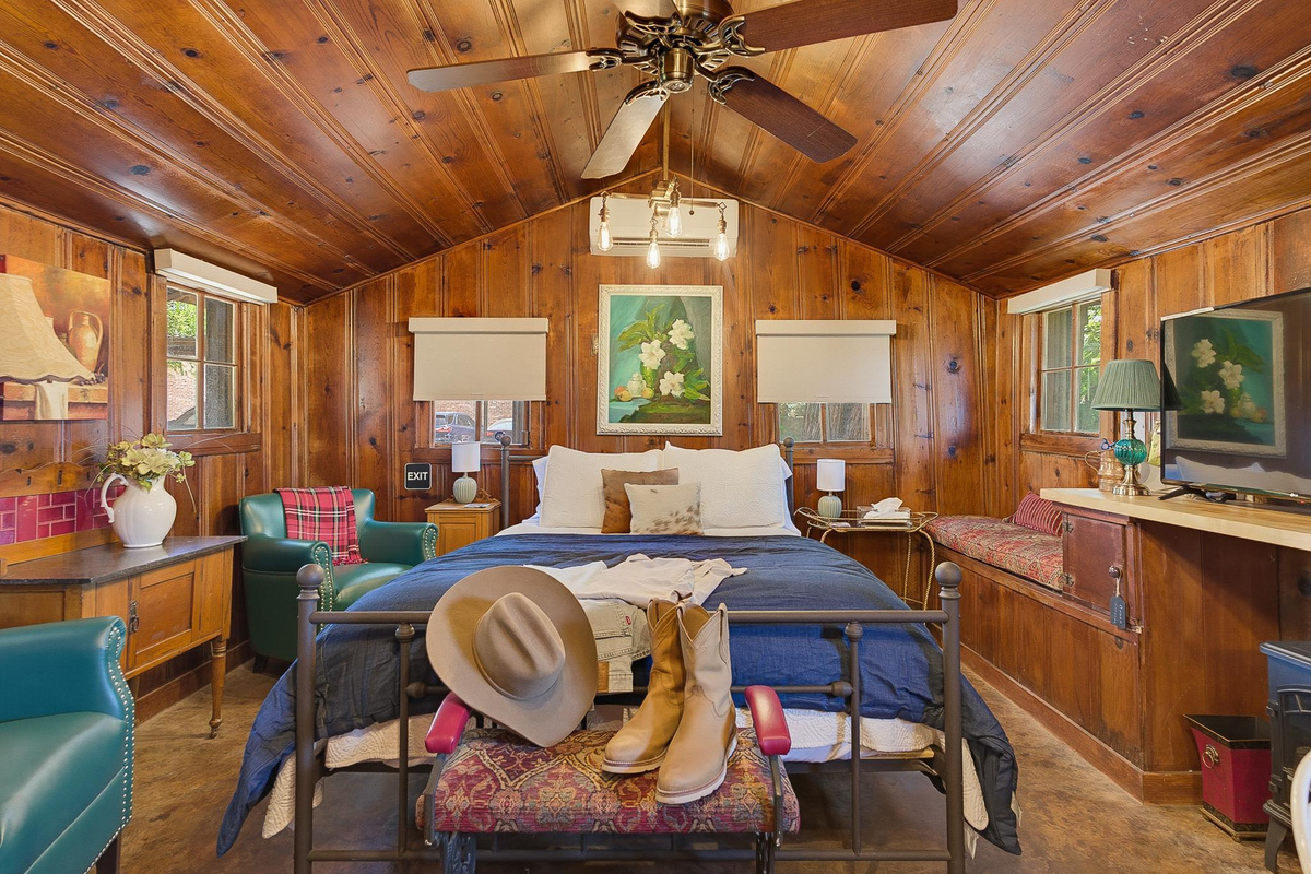 Papaw's Cabin rustic bedroom with wrought-iron queen bed, cowboy hat and boots, leather chairs, and warm wood paneling. Historic Waco, Texas log cabin escape.