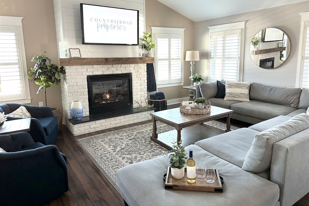 Gather in this sun-drenched living room featuring vaulted ceilings and a cozy white brick fireplace. The sprawling sectional and navy accent chairs offer plenty of space to relax and watch a movie after a long day at the beach.