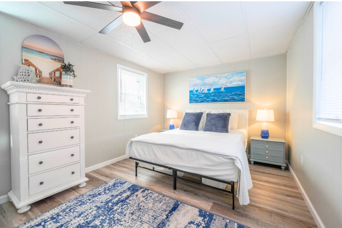 Bright bedroom with a comfortable bed, soft linens, and coastal artwork. Natural light, bedside lamps, and a ceiling fan create a calm space for restful nights, with storage furniture providing added convenience during your stay.