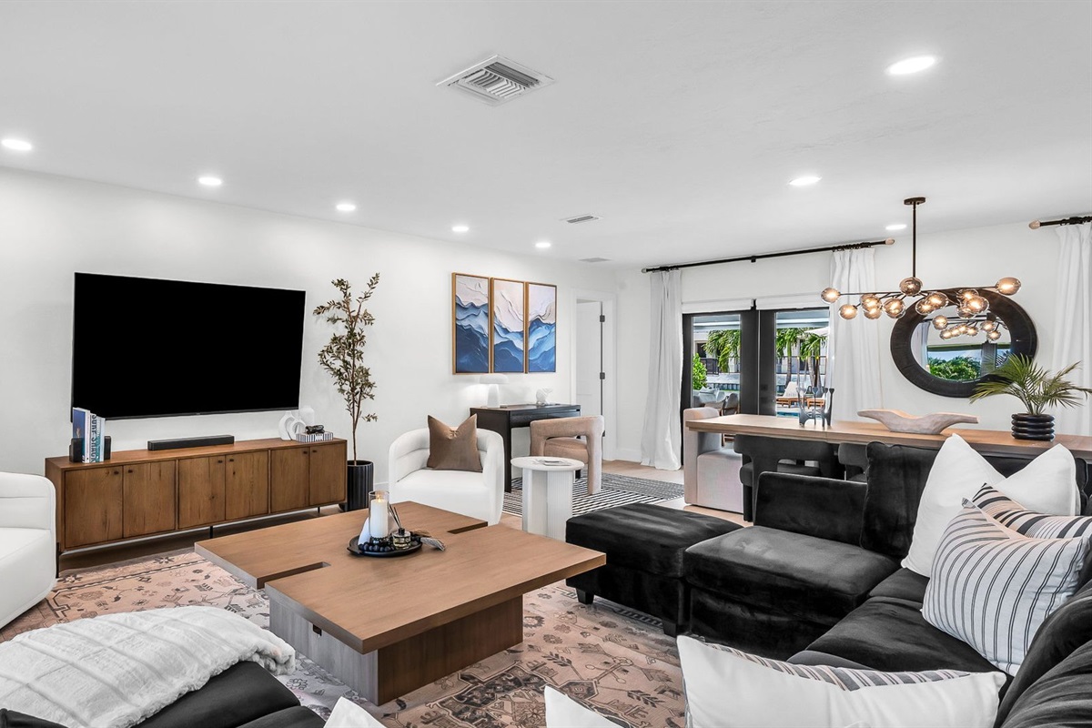Elegant open-concept living room featuring a plush black velvet sectional, 75” smart TV, and designer mid-century modern furnishings. Floor-to-ceiling windows and French doors fill the space with natural light and connect seamlessly to the outdoor pool.
