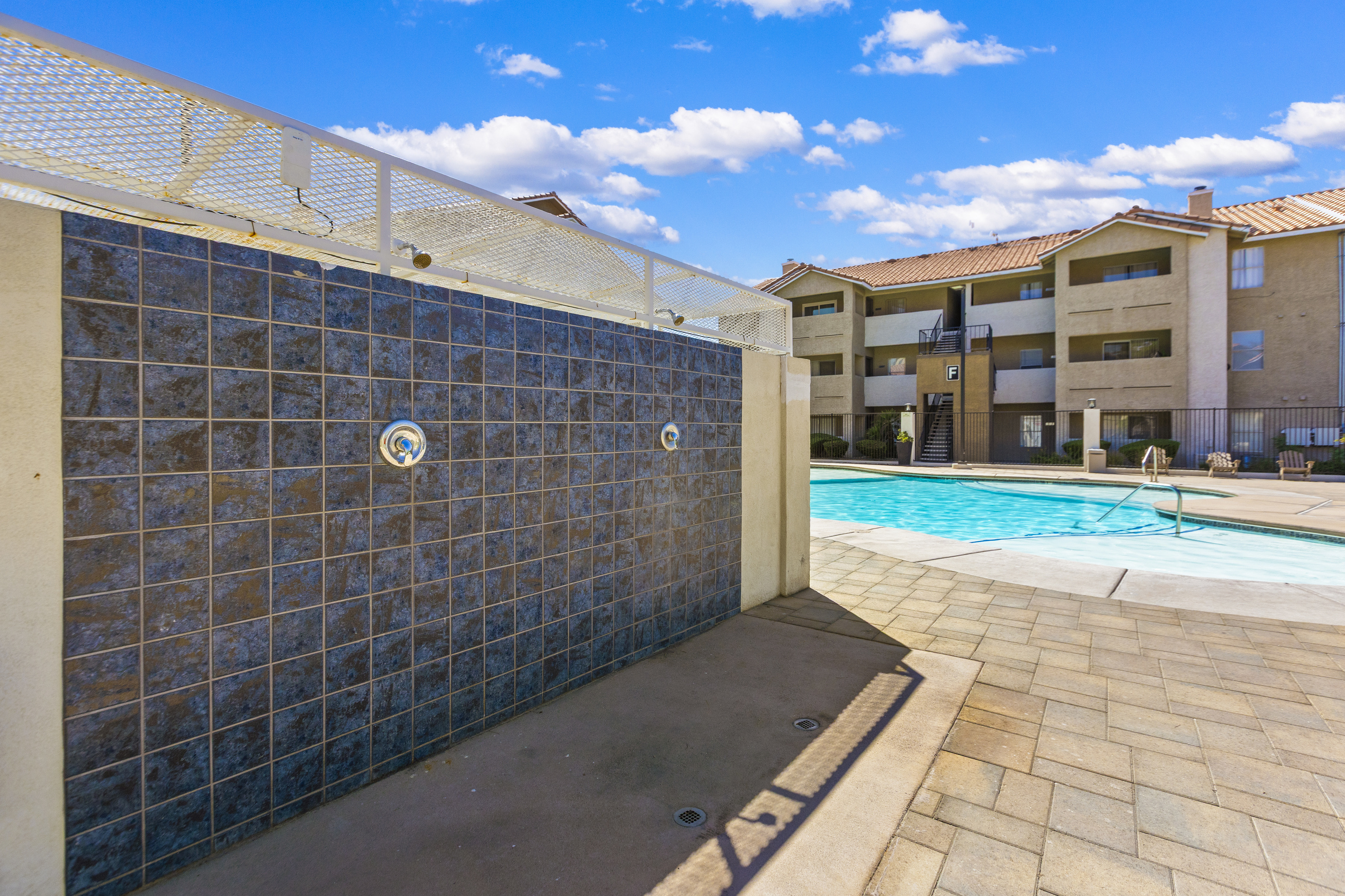 Shared pool area with outdoor shower station, paved pool deck, and apartment buildings nearby.
