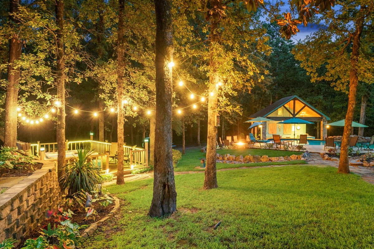 A dreamy nighttime retreat! This beautifully lit backyard features a sparkling pool, cozy fire pit, and a charming outdoor kitchen. Relax under the umbrellas or take a dip—perfect for unwinding in a serene setting.