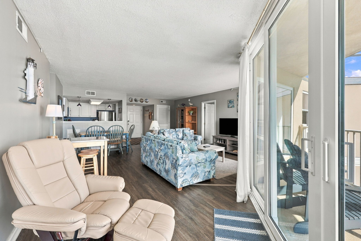 Comfortable living room featuring a neutral recliner chair, floral sofa, wood-look flooring, and sliding glass doors leading out to the oceanfront balcony.