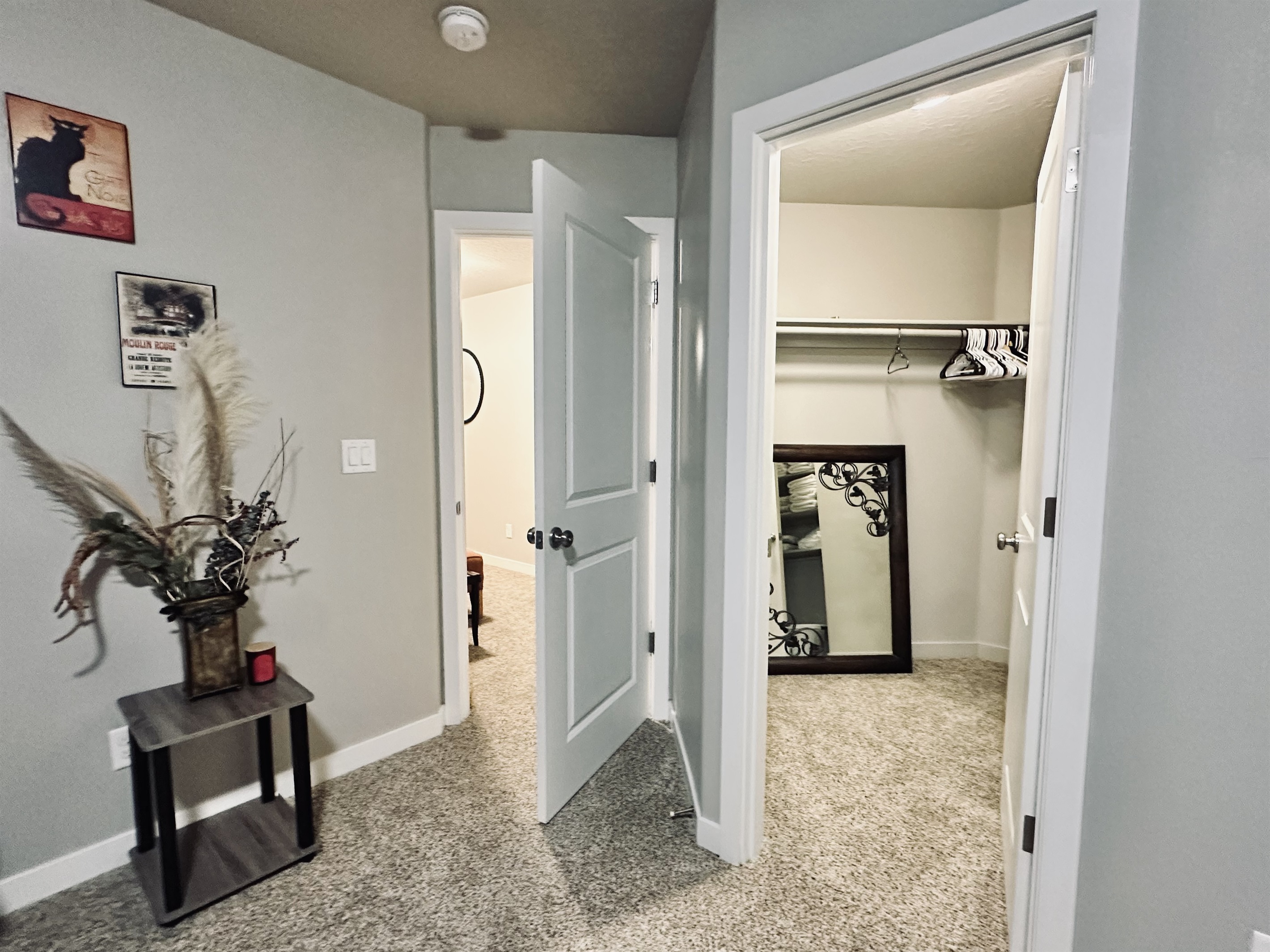 A light gray room features an open door leading to a small closet with hangers, a large mirror leaning against the wall, a side table with a decorative plant and a red candle, and a glimpse of another room in the background.