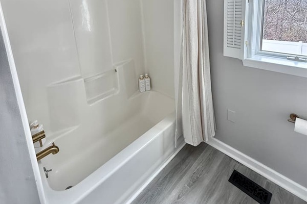 You will see a clean, modern bathroom space featuring a white tub-shower combination accented with elegant gold-tone fixtures. A light, neutral-toned shower curtain and shuttered window complete the serene, coastal aesthetic.