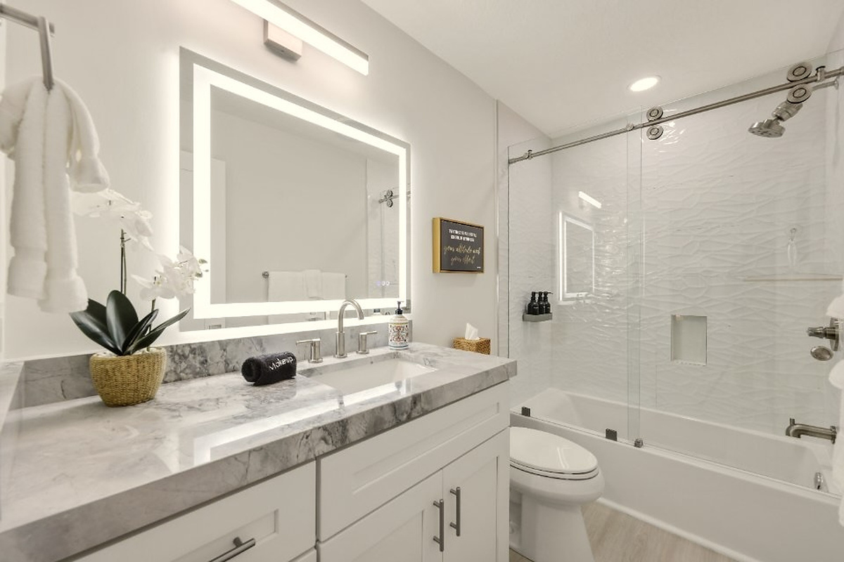 A bright full bathroom includes an illuminated mirror, modern vanity, and glass-enclosed tub/shower for easy daily routines.