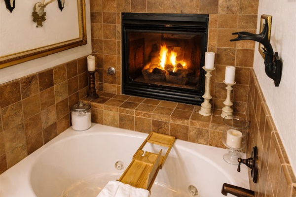 Luxuriate in a spa-like soaking tub beneath a glowing gas fireplace. This serene bathroom retreat features a deep jetted tub, warm stone tile, plush towels, and curated décor—perfect for unwinding after mountain adventures with a glass of wine and total re
