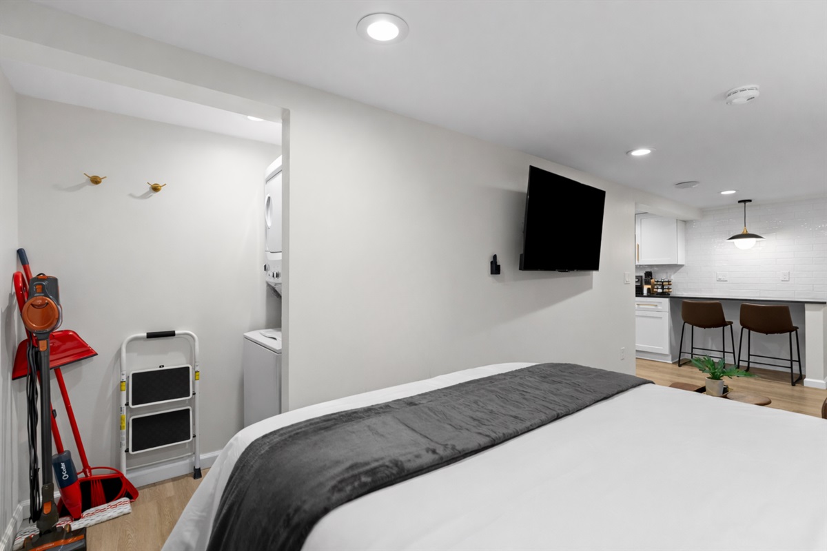 Comfortable sleeping area with a plush bed, wall-mounted smart TV, and an open layout that flows easily into the kitchen and living space.