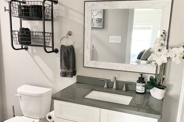Enjoy a modern and clean guest bathroom featuring a sleek grey quartz countertop and a white vanity. The space is thoughtfully designed with industrial-style wall baskets for storage, a textured frame mirror, and elegant orchid accents.