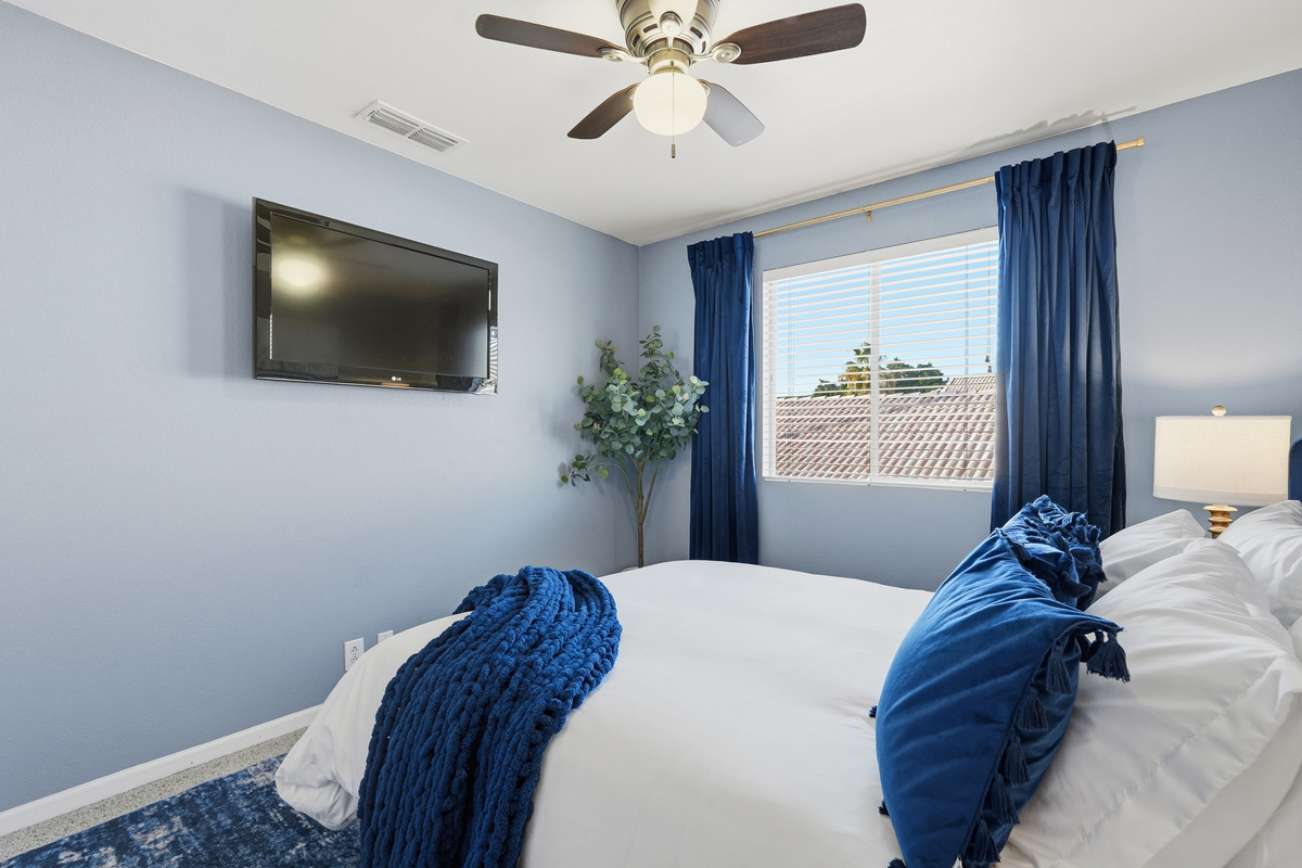 Bedroom with wall-mounted TV, ceiling fan, blue accent styling, and natural light for a comfortable stay.