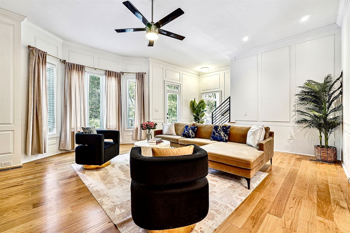 Situated next to the kitchen, this main-floor living room offers a warm, upscale ambiance with a brown L-shaped sectional, two black accent chairs, gold and black design accents, a smart TV, and a fireplace, perfect for entertaining or unwinding.