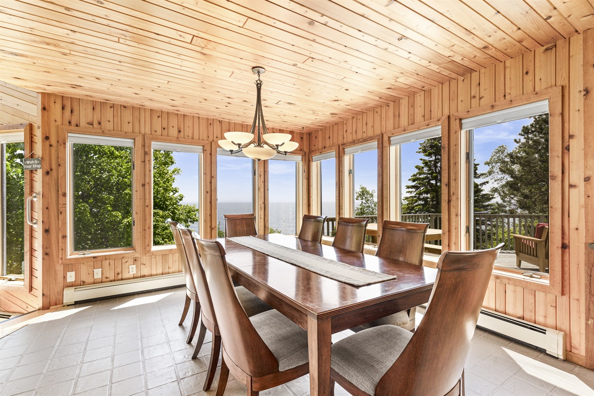 Bright dining room surrounded by windows, with a large table for shared meals, coffee, and conversation while taking in Lake Superior, shoreline, deck, and tree-filled views. A warm wood-paneled gathering space perfect for family dinners and game nights!
