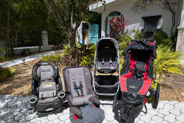 Lineup of complimentary family travel gear at Villa Coral including a Graco toddler car seat, infant car seat, booster seat, standard stroller, and Baby Jogger jogging stroller on a paved driveway in Dorado, Puerto Rico.