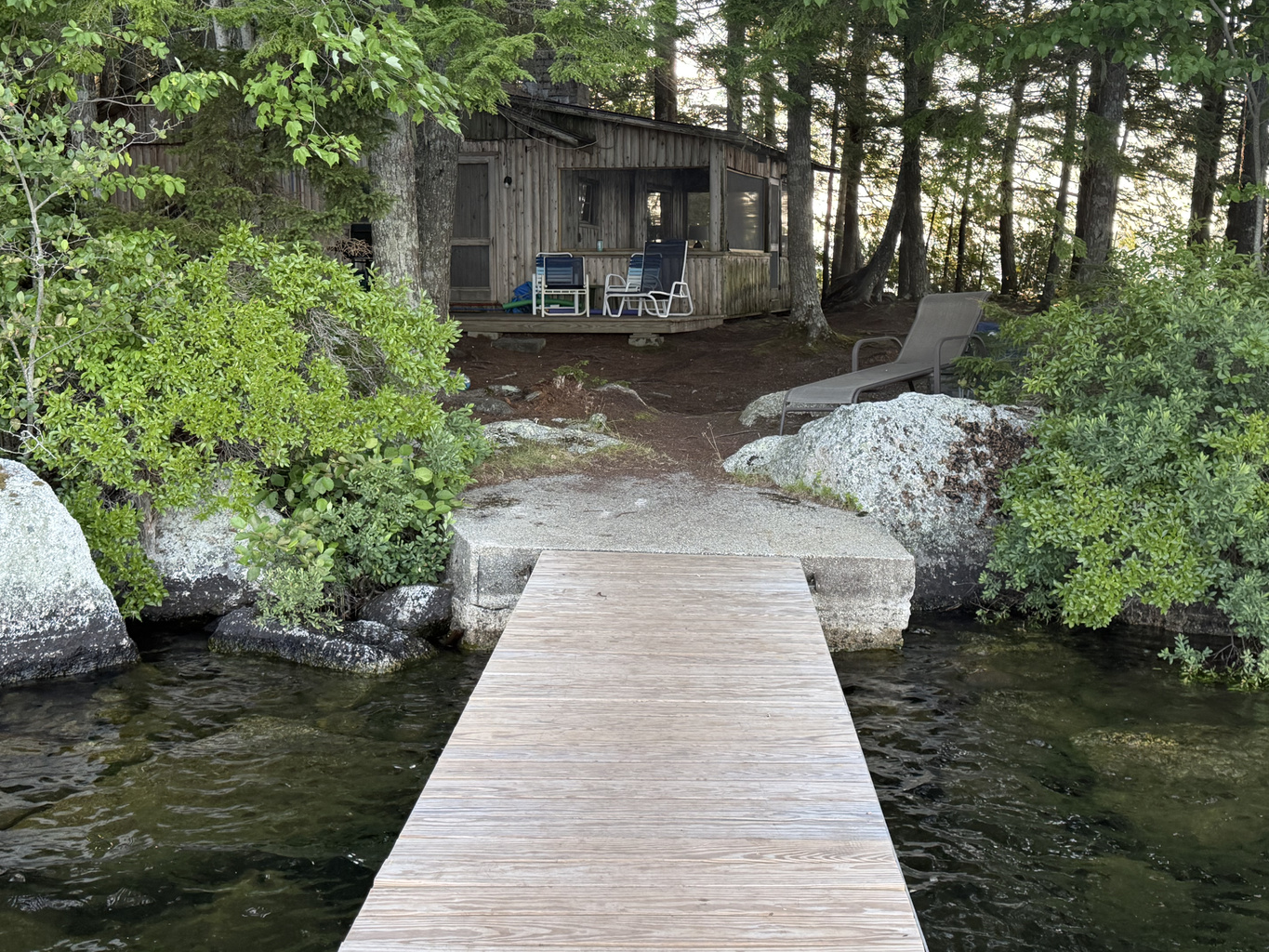 View looking back at camp standing on your private wooden dock! Steps off the concrete landing lead up natural granite rocks and pine paths to the deck and screened porch. Clear Beech Hill Pond water below—great spot to fish or launch kayaks!