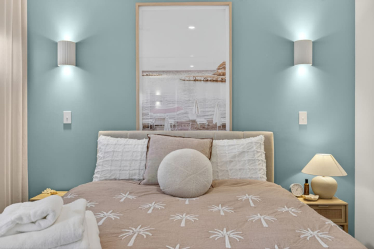 Calm coastal bedroom with a padded bedhead, palm-print linen, reading lights on both sides and fresh towels laid out ready.