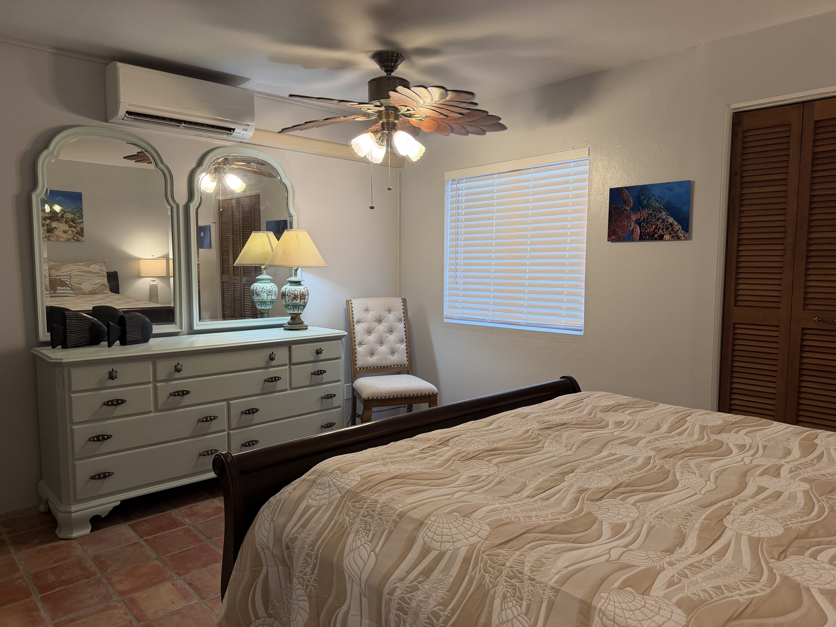 This bedroom offers a plush queen sleigh bed, classic Saltillo tile floors, and soft coastal accents. Complete with air conditioning, ceiling fan, dual nightstands with USB charging lamps, and a spacious double-mirror dresser.