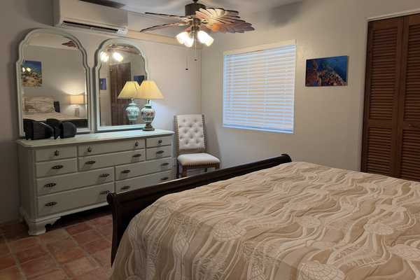 This bedroom offers a plush queen sleigh bed, classic Saltillo tile floors, and soft coastal accents. Complete with air conditioning, ceiling fan, dual nightstands with USB charging lamps, and a spacious double-mirror dresser.