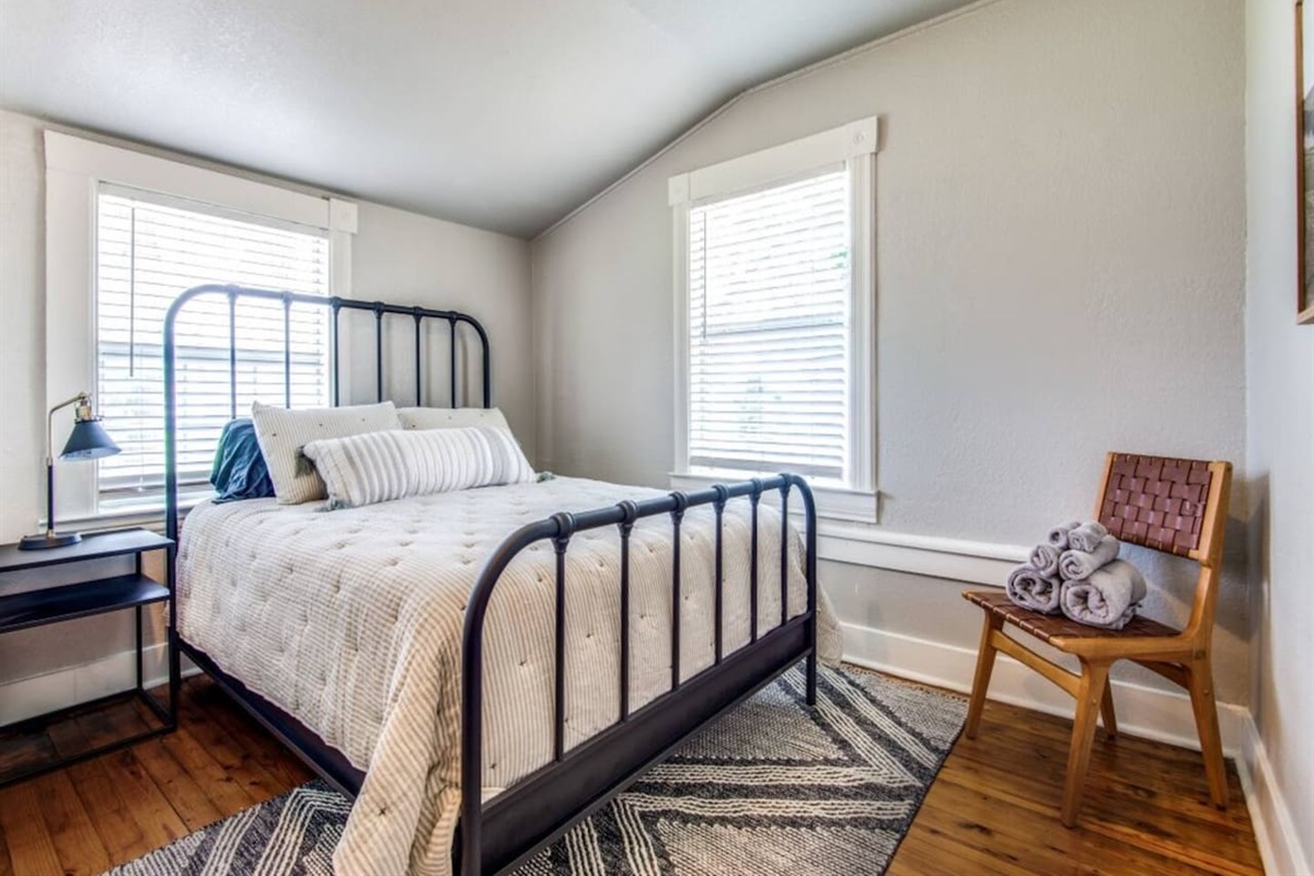 Light-filled queen bedroom with iron-frame headboard, woven rug, hardwood floors, and bright window. Sleeps 2 as part of a 3BR/2.5BA farmhouse sleeping up to 7 near The Milestone and The Grove wedding venues in Aubrey TX.