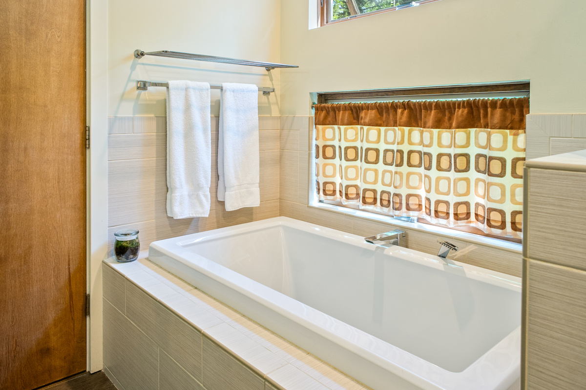Unwind in style at "The Crag." The primary en-suite features a deep, tile-surround soaking tub with modern chrome fixtures, plush towels, and cozy retro cafe curtains for the ultimate post-hike spa experience near Acadia.