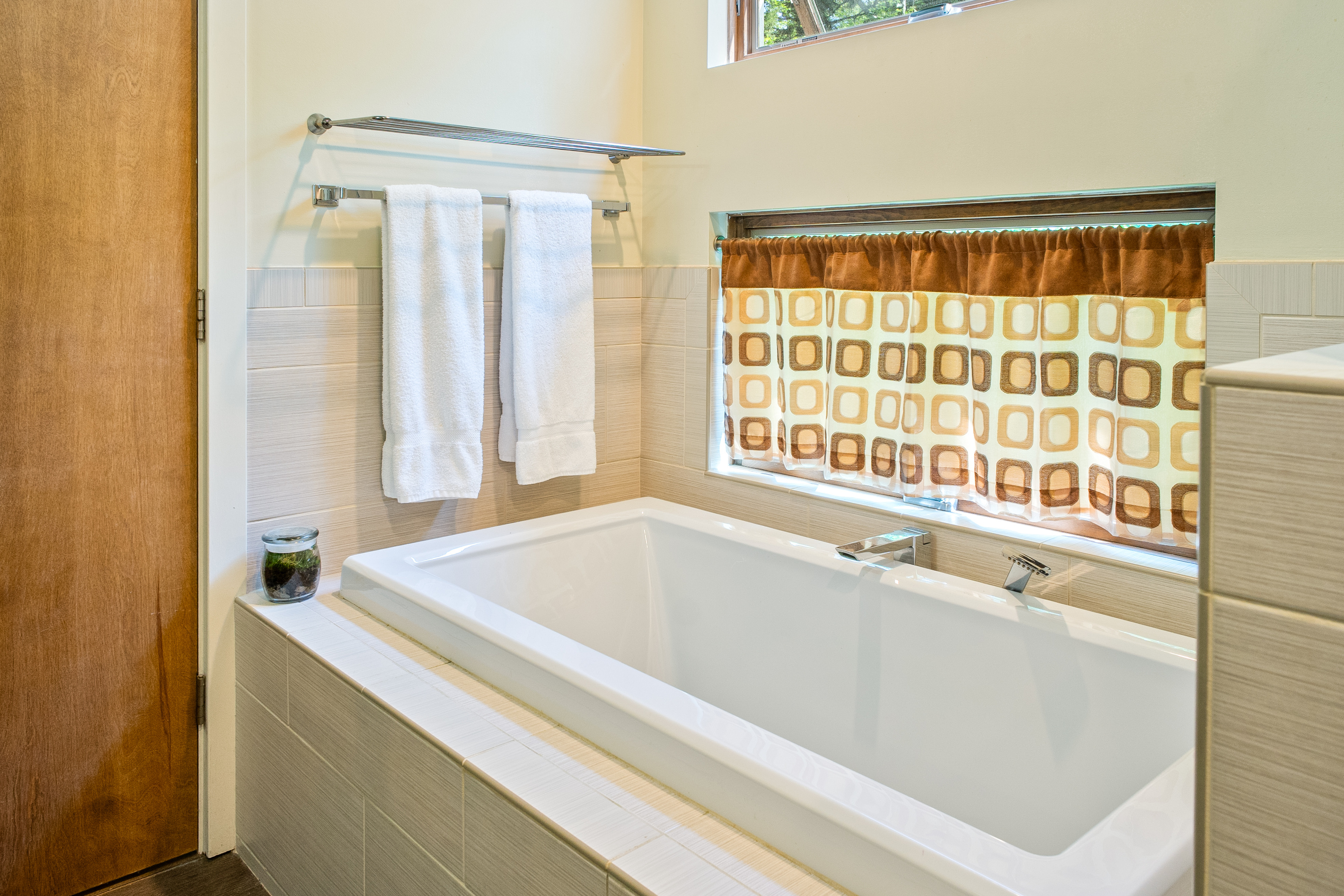 Unwind in style at "The Crag." The primary en-suite features a deep, tile-surround soaking tub with modern chrome fixtures, plush towels, and cozy retro cafe curtains for the ultimate post-hike spa experience near Acadia.