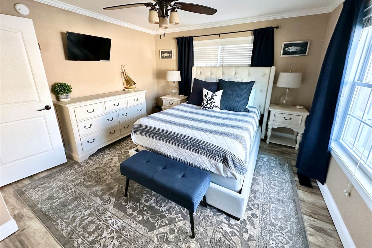 This nautical-inspired bedroom offers a tufted queen bed, a mounted TV, and a spacious dresser. A blue upholstered bench, patterned area rug, and coastal accents create a relaxing retreat for your stay.