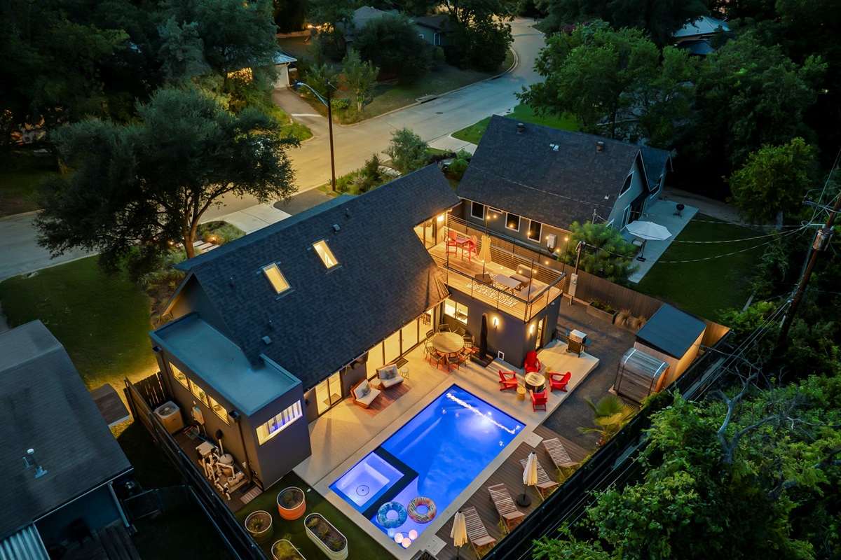 An aerial nighttime view of a modern home, showcasing a large swimming pool, a hot tub, and a multi-level deck for lounging and entertaining, all beautifully lit by string lights and outdoor fixtures.