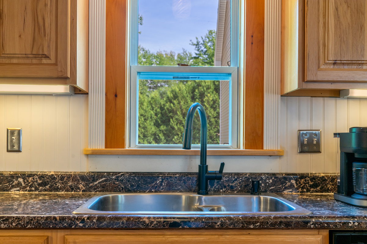 Prepare meals efficiently at the wide stainless steel kitchen sink featuring a modern matte black gooseneck faucet with a pull-down sprayer. The station is detailed with dark stone-patterned laminate countertops and a bright window view.