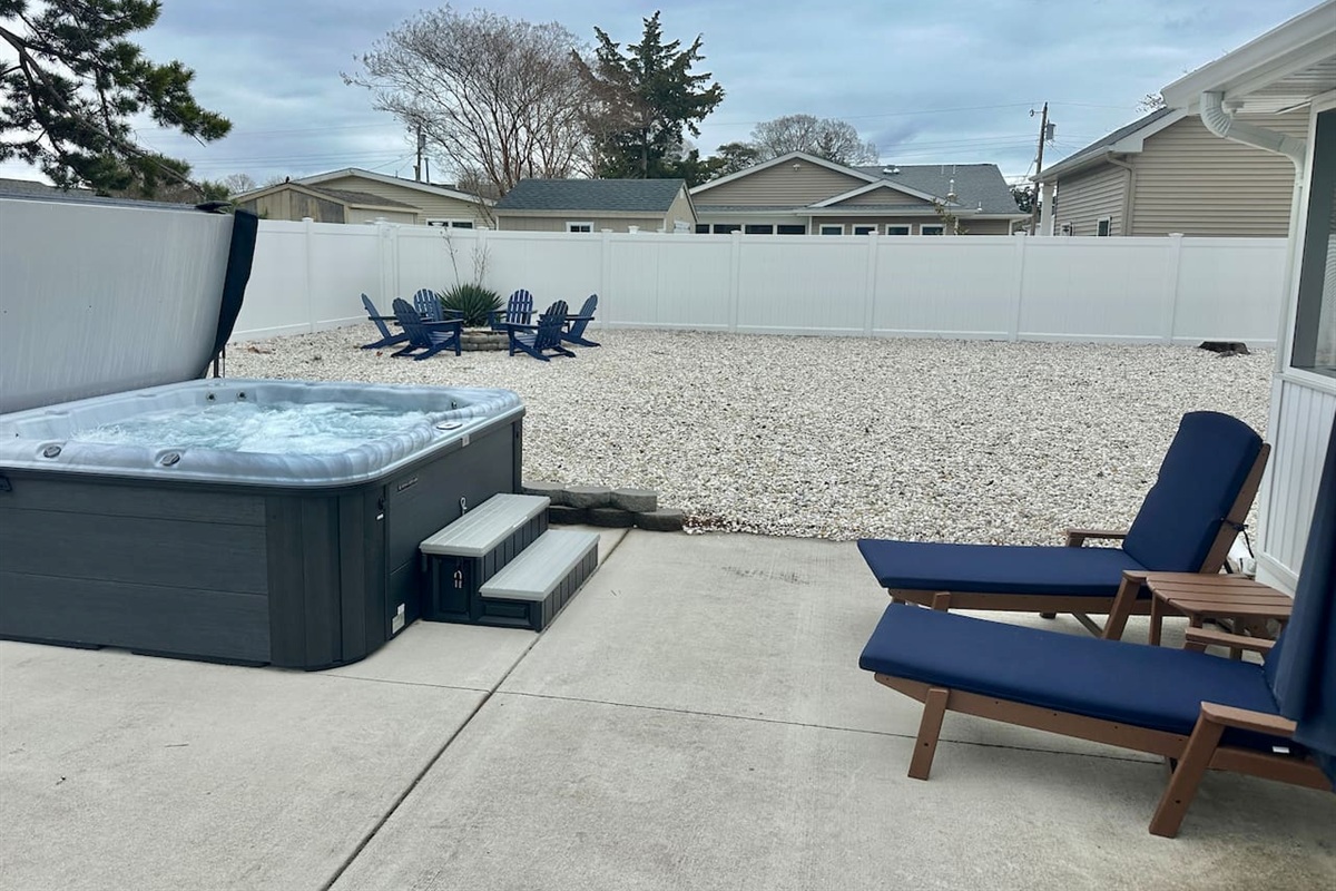 Unwind in your private outdoor retreat featuring a bubbling hot tub and comfortable lounge chairs with navy cushions. The spacious, fenced-in yard also includes a gravel area with Adirondack chairs gathered around a fire pit, perfect for starlit evenings.
