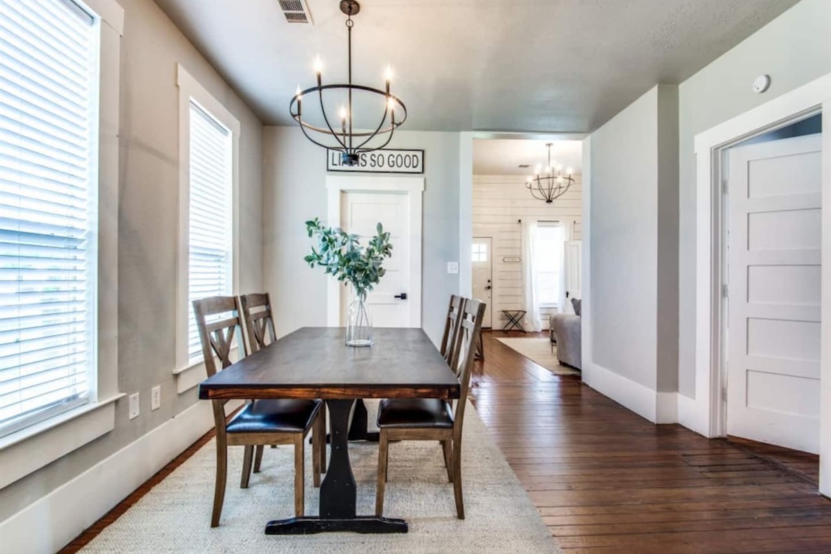 Farmhouse dining table with upholstered chairs, statement chandelier, shiplap accent wall, and hardwood floors. Bright natural light from large windows. Modern farmhouse Airbnb in downtown Aubrey TX steps from S Main Street dining.